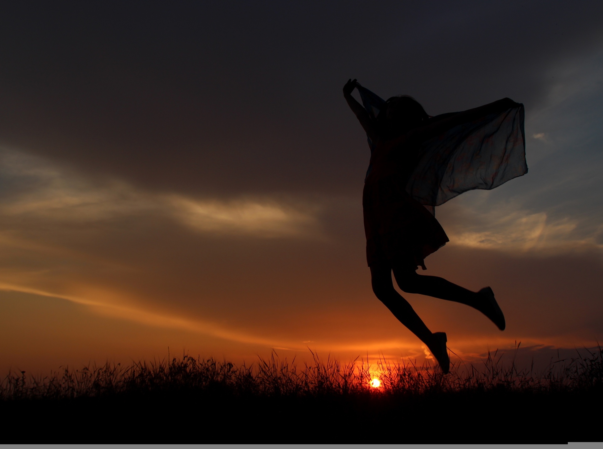 A woman leaping joyfully in an open field at sunset, arms outstretched with a scarf trailing behind her