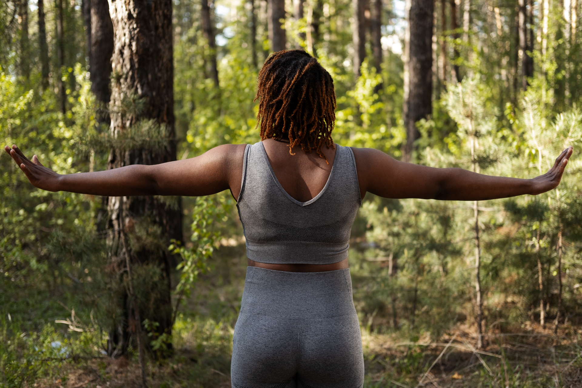 A Black woman standing in a sunlit forest with her arms extended wide during outdoor movement